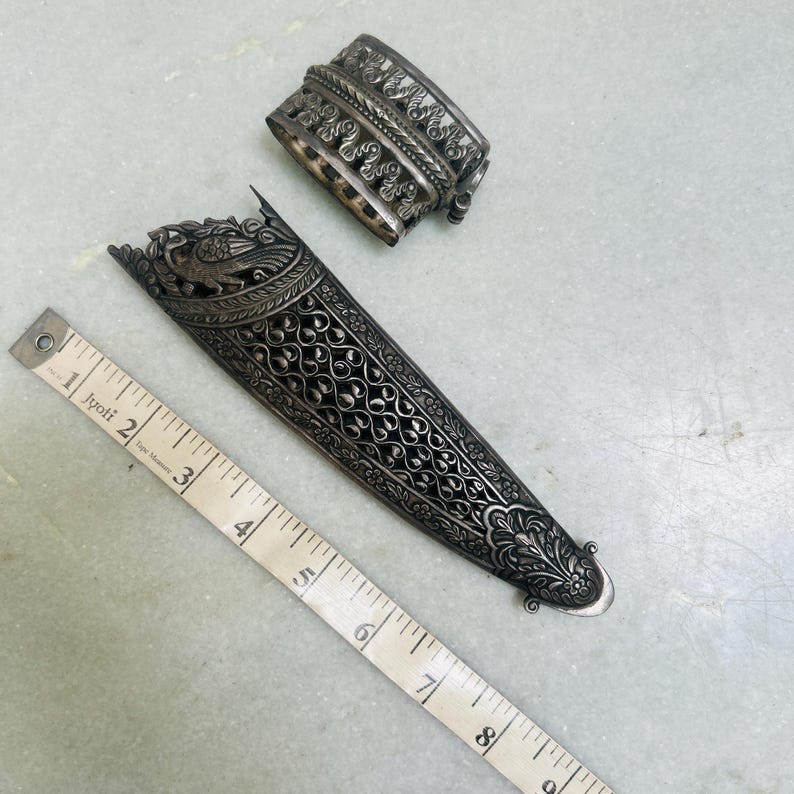 May include: A silver dagger sheath with intricate floral and bird designs. The sheath is approximately 7 inches long and has a decorative silver cap.