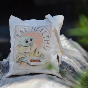 May include: A white canvas tote bag with a green character holding a box of fruit. The bag has the text "Eat Healthy" printed on it.