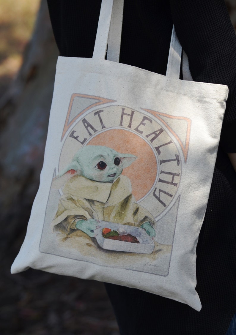 May include: A white canvas tote bag with a green character holding a container of food. The bag has the text "Eat Healthy" printed on it.