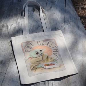 May include: A white canvas tote bag with a graphic of a green character eating a meal. The graphic is surrounded by a peach-colored circle with the text "Eat Healthy" in a vintage font.