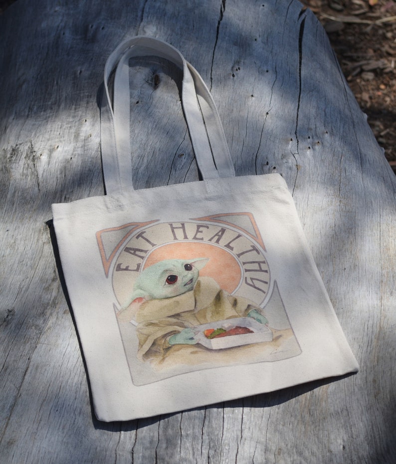 May include: A white canvas tote bag with a graphic of a green character eating a meal. The graphic is surrounded by a peach-colored circle with the text "Eat Healthy" in a vintage font.