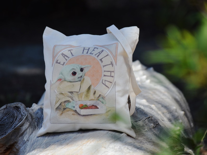 May include: A white canvas tote bag with a green character holding a box of fruit. The bag has the text "Eat Healthy" printed on it.