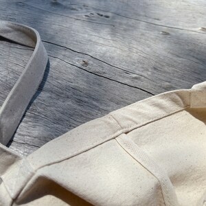 May include: A close-up of a natural-colored canvas tote bag with a single strap. The bag is folded in half and the strap is laying flat on a wooden surface.