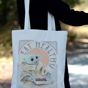May include: A white canvas tote bag with a graphic of a green alien character holding a container of food. The text "Eat Healthy" is written in a decorative font around the character.