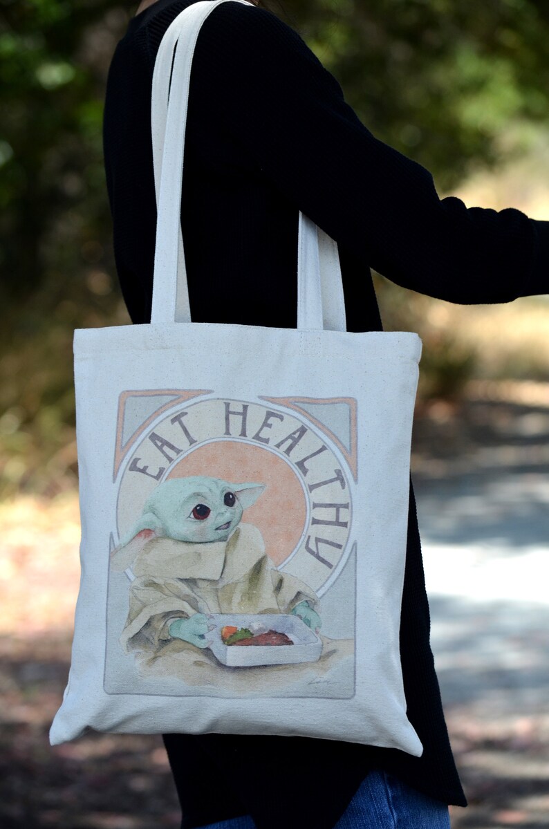 May include: A white canvas tote bag with a graphic of a green alien character holding a container of food. The text "Eat Healthy" is written in a decorative font around the character.