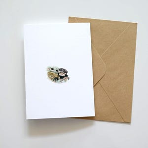 May include: A white greeting card with a small illustration of two characters, and a brown envelope. The card is blank, with the illustration in the center. The envelope is made of brown paper.