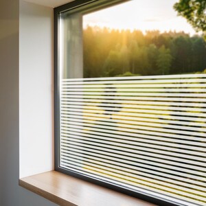 Puede incluir: Una ventana con una película de privacidad blanca mate con líneas horizontales. La película se aplica en el interior de la ventana. La ventana está enmarcada en negro y tiene un alféizar de madera.