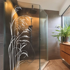 Può includere: Un bagno moderno con una porta doccia in vetro che presenta una silhouette bianca di erba alta. La doccia ha un soffione cromato e una maniglia argentata. Il bagno ha un pavimento in legno e un mobile in legno con cassetti.