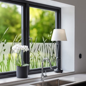 K&ouml;nnte beinhalten: Ein Fenster mit einer mattierten Glasfolie, die eine Silhouette von hohem Gras zeigt. Eine wei&szlig;e Orchideenpflanze steht auf dem Fensterbrett unter dem Fenster. Eine schwarze Lampe mit einem wei&szlig;en Schirm steht auf der Theke neben dem Fenster. Ein silberner Wasserhahn ist im Waschbecken zu sehen.