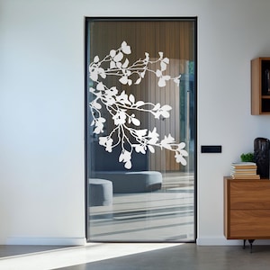 Può includere: Una porta in vetro smerigliato con una silhouette bianca di un ramo di magnolia con foglie e fiori. La porta è incorniciata in nero e ha una maniglia argentata.