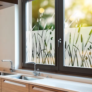 K&ouml;nnte beinhalten: Ein K&uuml;chenfenster mit einer Milchglas-Sichtschutzfolie, die eine Silhouette von hohem Gras zeigt. Das Fenster ist mit dunkelbraunem Holz umrahmt und hat darunter eine wei&szlig;e Arbeitsplatte. Zwei verchromte Wasserh&auml;hne sind in der Sp&uuml;le sichtbar.