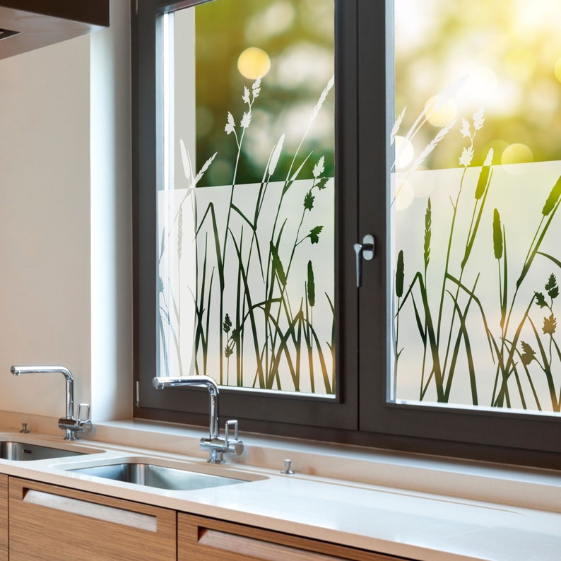 K&ouml;nnte beinhalten: Ein K&uuml;chenfenster mit einer Milchglas-Sichtschutzfolie, die eine Silhouette von hohem Gras zeigt. Das Fenster ist mit dunkelbraunem Holz umrahmt und hat darunter eine wei&szlig;e Arbeitsplatte. Zwei verchromte Wasserh&auml;hne sind in der Sp&uuml;le sichtbar.
