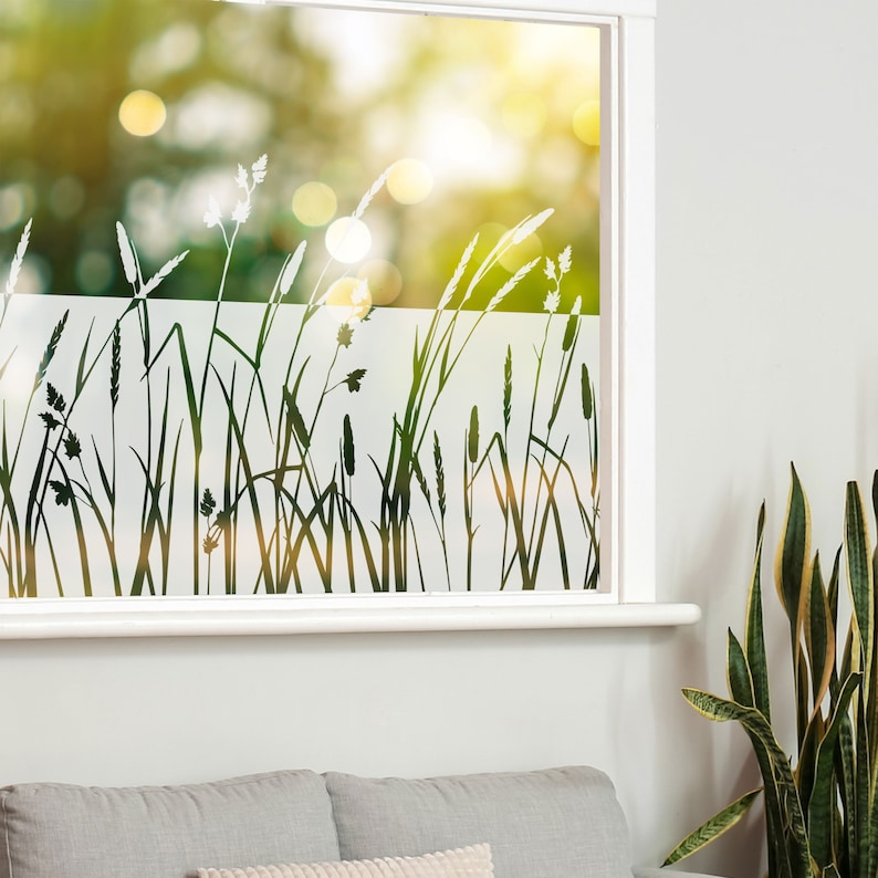 K&ouml;nnte beinhalten: Ein Fenster mit einer mattierten Glasfolie, die eine Silhouette von hohem Gras und Blumen zeigt. Die Folie ist hellgrau und das Gras und die Blumen sind dunkelgrau. Das Fenster ist wei&szlig; umrahmt.