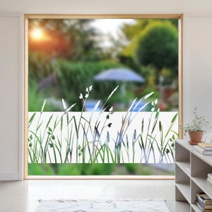 K&ouml;nnte beinhalten: Eine matte Fensterfolie mit einem Silhouetten-Design aus hohen Gr&auml;sern und Wildblumen. Die Folie ist auf ein Fenster mit einem verschwommenen Blick auf einen Garten im Hintergrund angebracht.