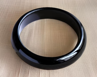 May include: A black bangle bracelet made of polished resin. The bracelet is smooth and shiny.