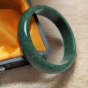 May include: A green jade bangle bracelet with a smooth, polished finish. The bracelet is displayed in a yellow velvet box.