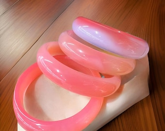 Peut inclure: Quatre bracelets translucides aux tons roses et violets. Les bracelets sont empilés, avec des couleurs allant du rose foncé au violet clair. Ils sont faits d'un matériau lisse et brillant. Un accessoire élégant.