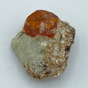 Puede incluir: Una pequeña gema de color rojo anaranjado rugosa rodeada de cristales blancos y marrones.