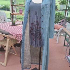 May include: A long-sleeved light blue denim duster coat with a button-down front, worn over a grey sleeveless dress featuring a dark red floral graphic print on the front and a ruffled hemline. The interior of the denim coat has a beige floral lining.
