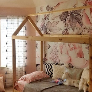 Op de afbeelding: Een houten bedframe in de vorm van een huis met een roze bloemenbehang erachter. Het bed is gemaakt met een grijs deken, roze en witte kussens en een paar knuffels.