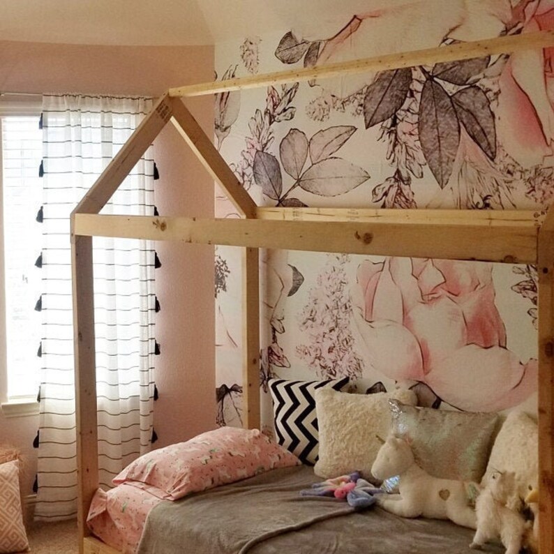 Op de afbeelding: Een houten bedframe in de vorm van een huis met een roze bloemenbehang erachter. Het bed is gemaakt met een grijs deken, roze en witte kussens en een paar knuffels.