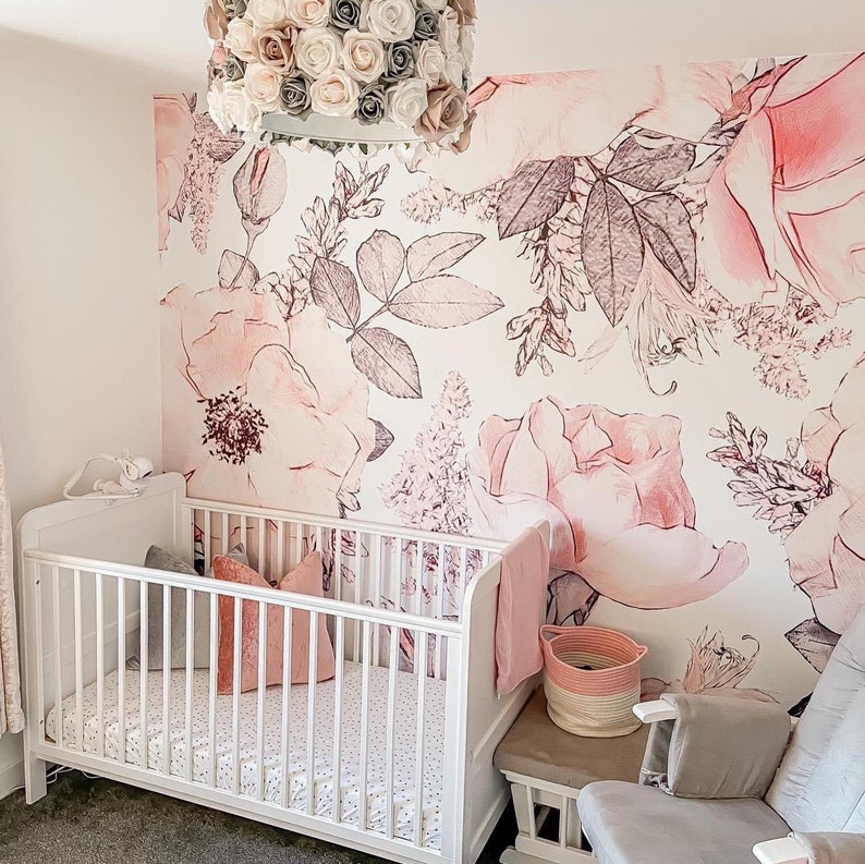 Op de afbeelding: Een wit wiegje met een roze bloemenbehang met een grote roze roos en een witte bloemenkrans die aan het plafond hangt.