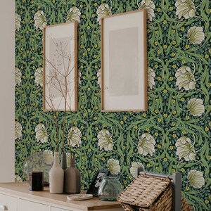 May include: A white dresser with three drawers and two doors, featuring a green floral wallpaper with white flowers and yellow accents. The dresser is decorated with two framed pictures, a vase with dried branches, and a glass jar. A wicker basket sits on a gray metal stand.