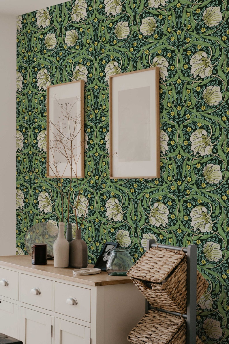 May include: A white dresser with three drawers and two doors, featuring a green floral wallpaper with white flowers and yellow accents. The dresser is decorated with two framed pictures, a vase with dried branches, and a glass jar. A wicker basket sits on a gray metal stand.