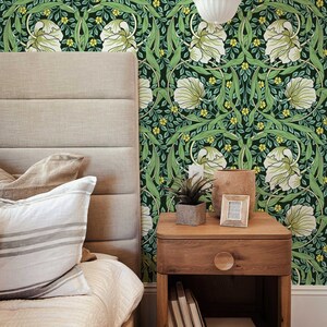 May include: A bedroom with a bed, a nightstand, and a green floral wallpaper. The nightstand has a drawer, a shelf, and a small plant on top. The bed has a white comforter and two pillows.