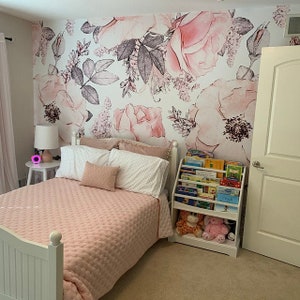 Op de afbeelding: Een wit bedframe met een roze gestikt dekbed en witte kussens. Het bed staat in een kamer met een bloemenbehang en een witte boekenkast gevuld met kinderboeken.