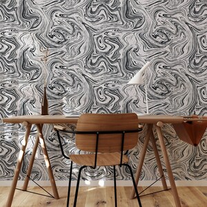 Op de afbeelding: Een houten bureau met een zwarte metalen stoel ervoor. Het bureau heeft een lichte houten afwerking en de stoel heeft een zwart metalen frame en een houten zitting. De muur achter het bureau is bedekt met zwart-wit marmerpapier.