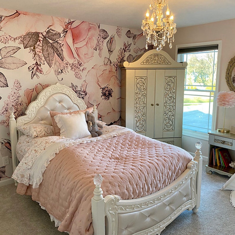 Op de afbeelding: Een wit, versierd bedframe met een roze, gestikte deken en behang met bloemenmotief. Het bed is versierd met kussens en een knuffeldier.
