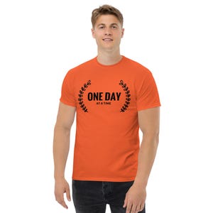 Puede incluir: Camiseta naranja con el texto negro "ONE DAY at a time", enmarcado por una corona de laurel negra. La camiseta es de manga corta y cuello redondo.