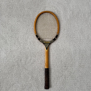 May include: Vintage wooden tennis racket with a light brown frame and a dark brown grip. The racket head features a woven string pattern. The handle has the word "Davis" printed on it. The racket is set against a gray background.