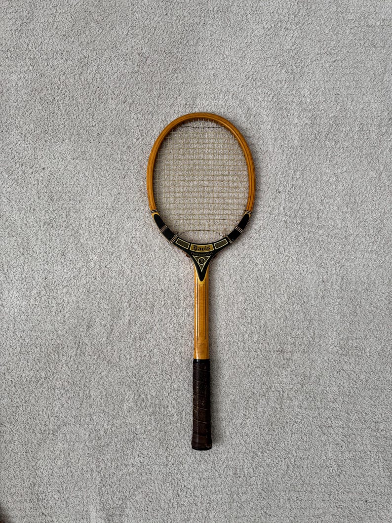 May include: Vintage wooden tennis racket with a light brown frame and a dark brown grip. The racket head features a woven string pattern. The handle has the word "Davis" printed on it. The racket is set against a gray background.