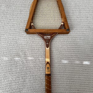 May include: Vintage wooden tennis racket with a brown handle and frame. The racket head is rectangular with a white mesh. The text "ULTRA Super Quality" is visible on the racket's throat. The grip is wrapped in brown leather.