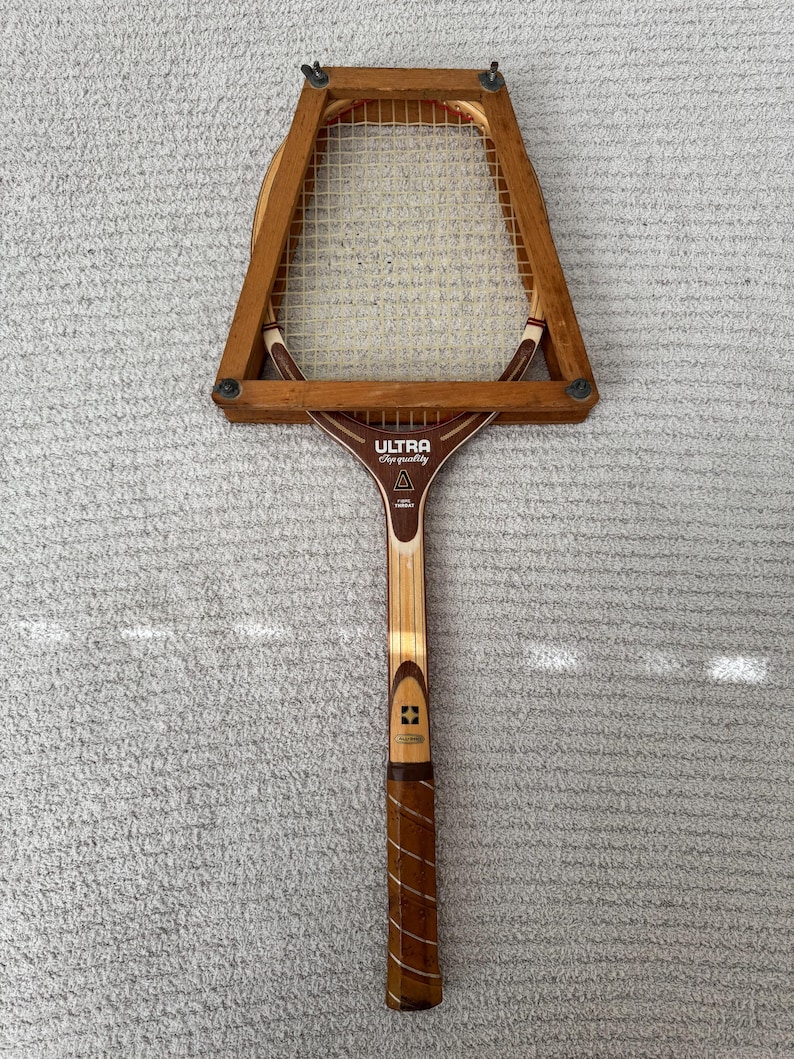 May include: Vintage wooden tennis racket with a brown handle and frame. The racket head is rectangular with a white mesh. The text "ULTRA Super Quality" is visible on the racket's throat. The grip is wrapped in brown leather.