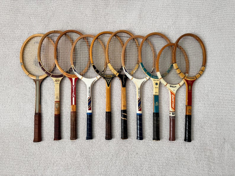 May include: A collection of vintage wooden tennis rackets with various handle and head designs. The rackets feature natural wood tones, with some having colored accents in red, blue, and yellow. The rackets are arranged on a light gray surface.