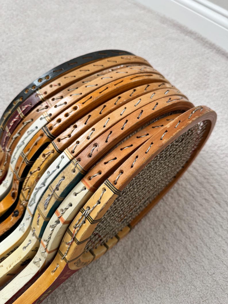 May include: A stack of vintage wooden tennis rackets. The rackets have natural wood tones, with some featuring painted accents in colors like black, white, and burgundy. The strings are visible, and the rackets appear to be in good condition.