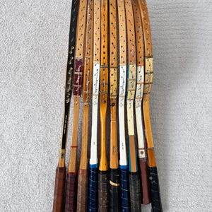 May include: A collection of vintage wooden tennis rackets. The rackets have brown, black, and white accents, with leather grips. The rackets are arranged in a fan shape, showcasing their unique designs and wear.