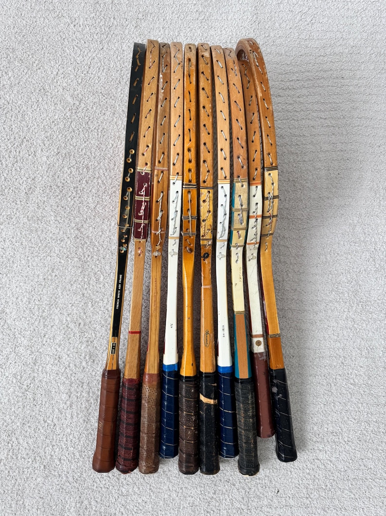 May include: A collection of vintage wooden tennis rackets. The rackets have brown, black, and white accents, with leather grips. The rackets are arranged in a fan shape, showcasing their unique designs and wear.