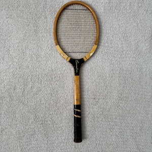 May include: Vintage wooden tennis racket with a light brown frame and black grip. The racket has a black and gold accent at the base of the head and the word "Coronation" is printed on the handle.