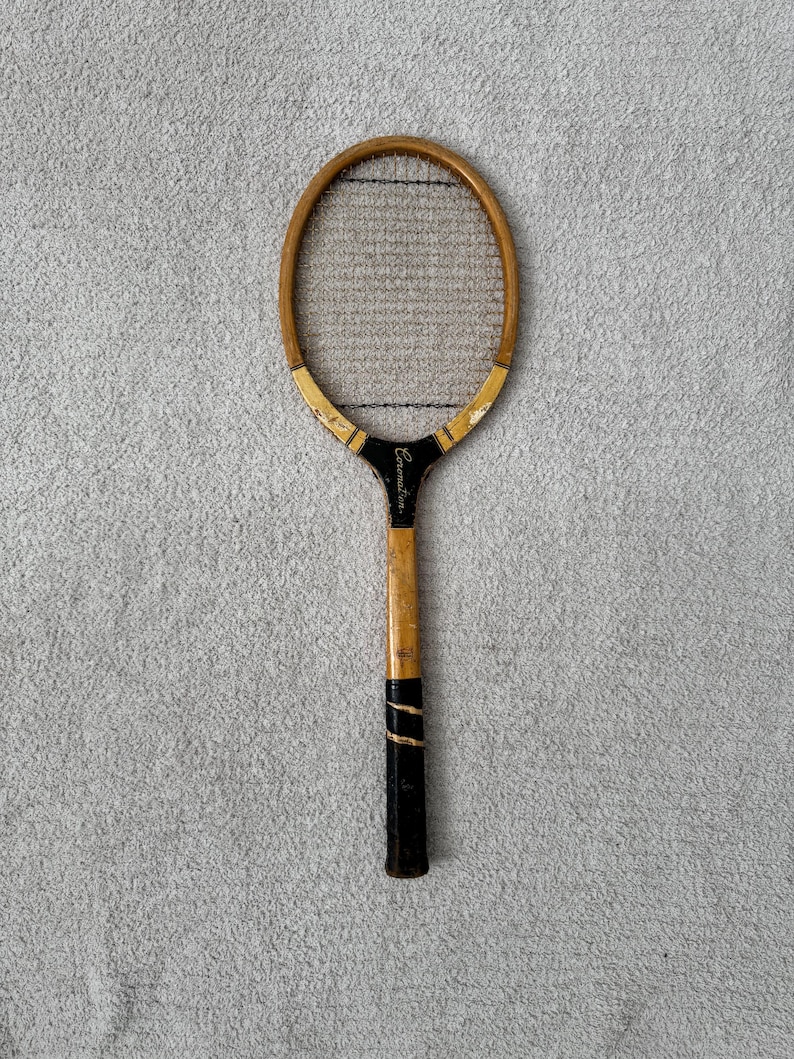 May include: Vintage wooden tennis racket with a light brown frame and black grip. The racket has a black and gold accent at the base of the head and the word "Coronation" is printed on the handle.