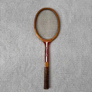 May include: Vintage Spalding tennis racket with a wooden frame and a red and white handle. The racket is labeled "Pancho Gonzales" and features a brown grip. The strings are visible within the oval frame.