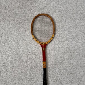 May include: Vintage Spalding tennis racket with a wooden frame and a red and black handle. The racket head is oval-shaped with a mesh stringing. The handle has a black grip and the brand name "Spalding" is visible.