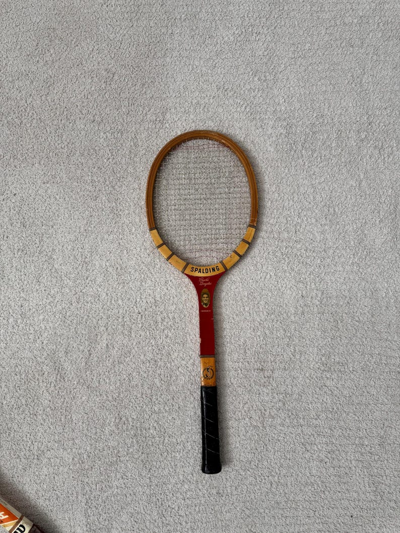 May include: Vintage Spalding tennis racket with a wooden frame and a red and black handle. The racket head is oval-shaped with a mesh stringing. The handle has a black grip and the brand name "Spalding" is visible.