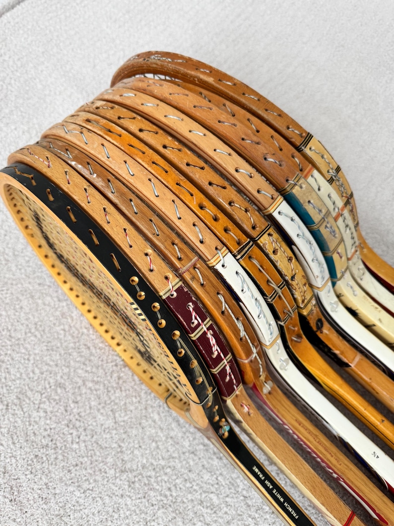 May include: A collection of vintage wooden tennis rackets. The rackets have natural wood frames with visible stringing and decorative accents in black, white, and burgundy. The rackets are stacked, showcasing their unique designs and aged appearance.