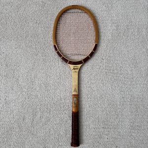 May include: Vintage wooden tennis racket with a light brown frame and a dark red grip. The racket has a light-colored handle with a portrait and the word "Jaguar" printed on the head.