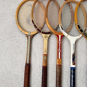 May include: A collection of vintage wooden tennis rackets. The rackets have brown leather grips, natural wood frames, and white netting. Some rackets have brand names like "Spalding" and "Junior Pro" printed on them. The rackets are arranged in a row.