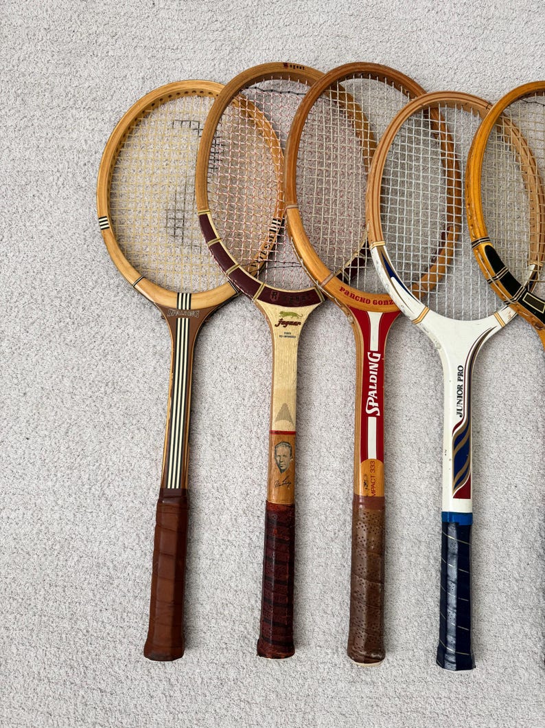 May include: A collection of vintage wooden tennis rackets. The rackets have brown leather grips, natural wood frames, and white netting. Some rackets have brand names like "Spalding" and "Junior Pro" printed on them. The rackets are arranged in a row.